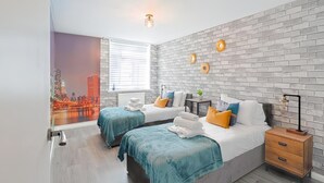 Ein Hotelzimmer mit einer Ziegelwand, zwei Betten mit türkisblau gestreuter Bettwäsche, einem Nachttisch und einem Wandgemälde mit Stadtansicht.