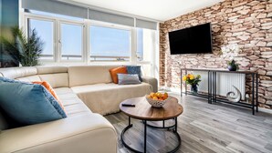 A modern living room with a stone wall, a flat-screen TV, a sofa with cushions, a coffee table with fruit, and large windows. - Central Brighton 1-bedroom Flat Modern & Bright Walk to Beach Sleeps 2 (Brighton)