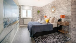 A bedroom with a brick wall, a bed with a grey blanket, two bedside lamps, and a patterned rug. - Central Brighton 1-bedroom Flat Modern & Bright Walk to Beach Sleeps 2 (Brighton)