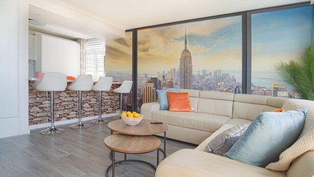 A modern living room with a large window, a stone accent wall, a beige sofa, and a small round table with a basket of oranges. - Central Brighton 1-bedroom Flat Modern & Bright Walk to Beach Sleeps 2 (Brighton)