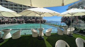 A poolside area with white wicker chairs and umbrellas, surrounded by a pool and a view of the sea.