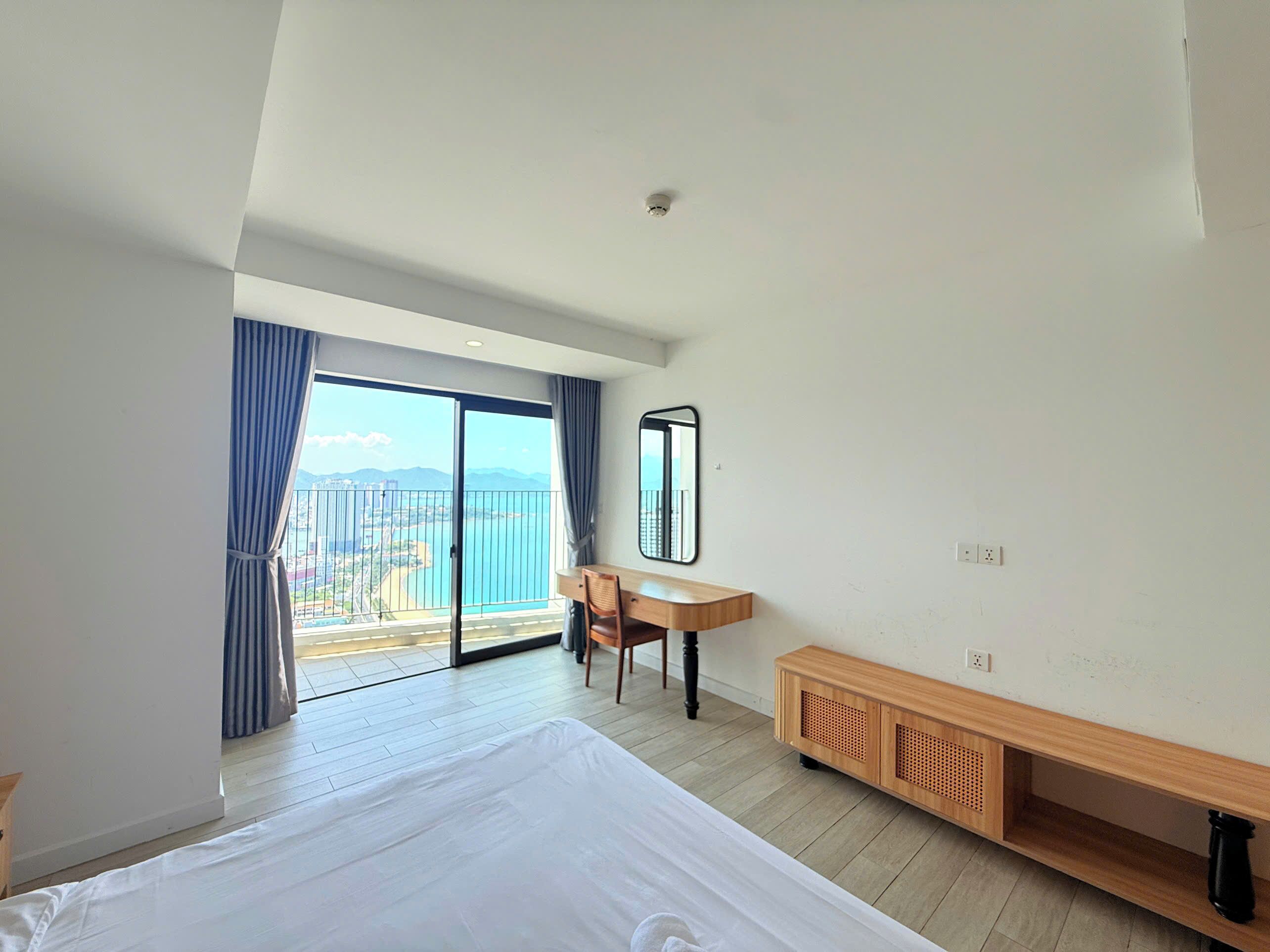 A hotel room with a bed, a desk, a chair, a mirror, and a view of a cityscape through a balcony.