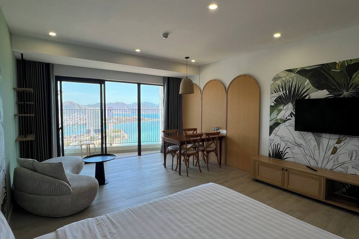 A modern hotel room with a large bed, a dining area with chairs, a TV mounted on the wall, and a balcony with a view of the city.