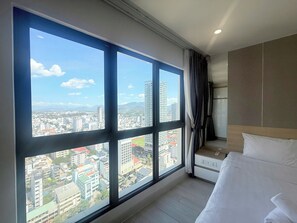 A hotel room with a large window, a bed, a nightstand, and a view of a cityscape.