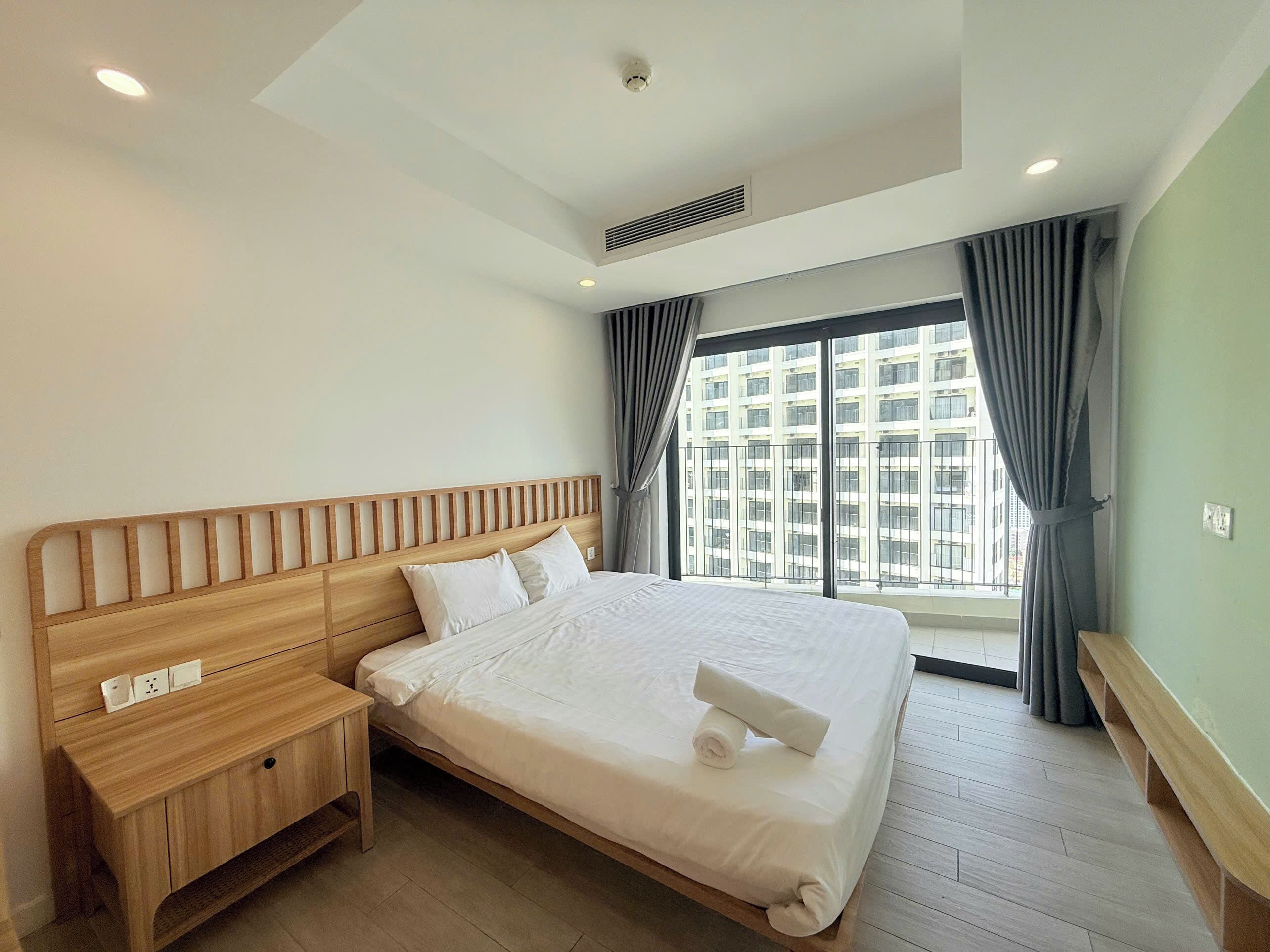 A hotel room with a large bed, a wooden nightstand, a window with curtains, and a view of a building.