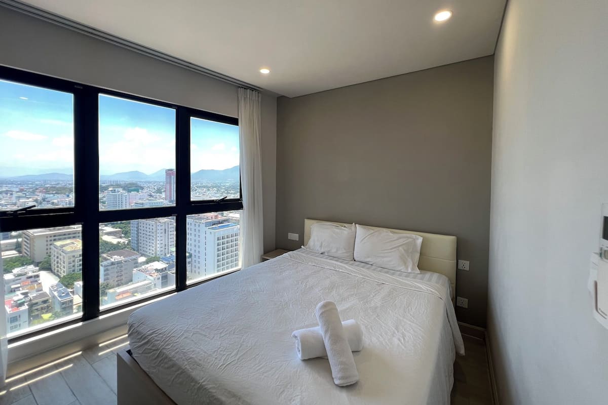 A modern hotel room with a large bed, white linens, and a city view through floor-to-ceiling windows.