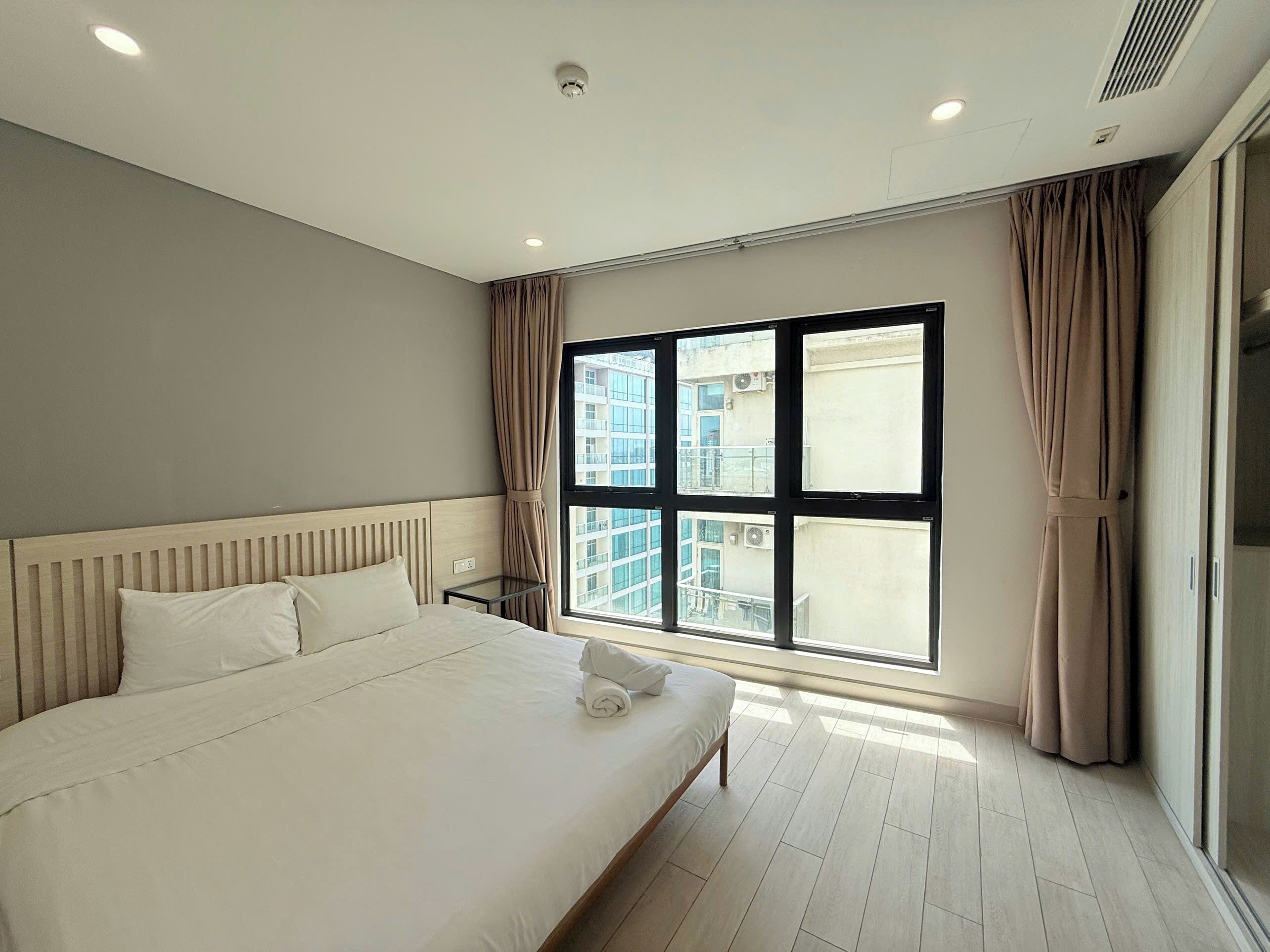 A hotel room with a large bed, a bedside table, a window with curtains, and a view of buildings outside.