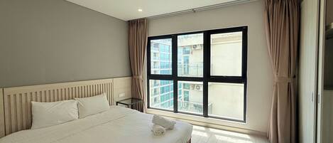 A hotel room with a large bed, a bedside table, a window with curtains, and a view of buildings outside.