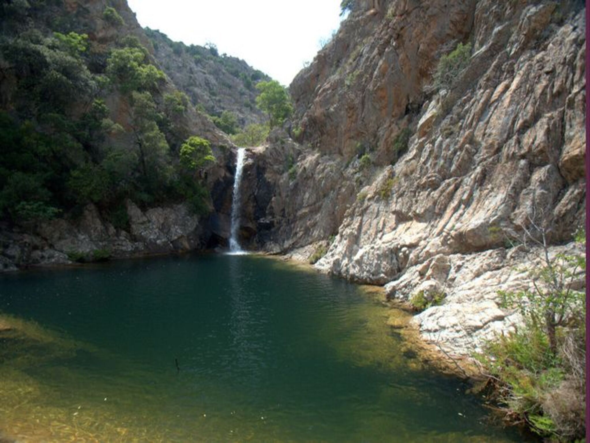 Eine natürliche Landschaft mit einem Wasserfall und einem klaren Wasserbecken, umgeben von Felswänden und viel Grün.