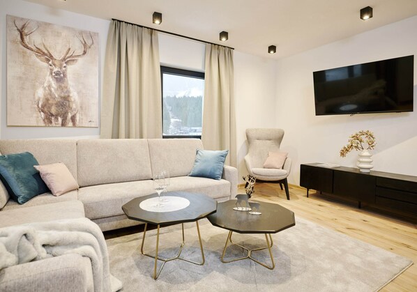 A modern living room with a sofa, two round tables, a TV, and a painting of a deer. - Apartment in Berwang With Ski-in & Sauna (Berwang)
