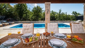 A wooden table set for breakfast with plates, glasses, and a fruit tray, overlooking a swimming pool. - **Villa Admiral: Your Idyllic Retreat in rural area of Trogir hinterland** (Blizna Gornja)