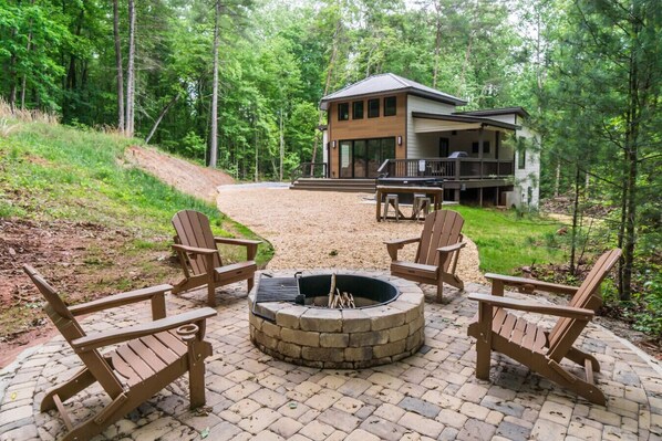 A cabin with a fire pit and four Adirondack chairs in a forest setting.