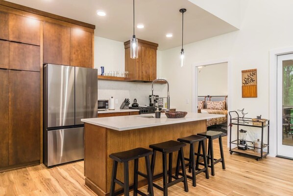 A modern kitchen with wooden cabinets, a stainless steel refrigerator, a central island with stools, and a dining area with a view of the outdoors.