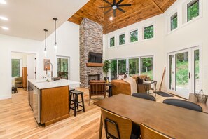 A spacious kitchen with a central island, a stone fireplace, and large windows offering a view of the outdoors.