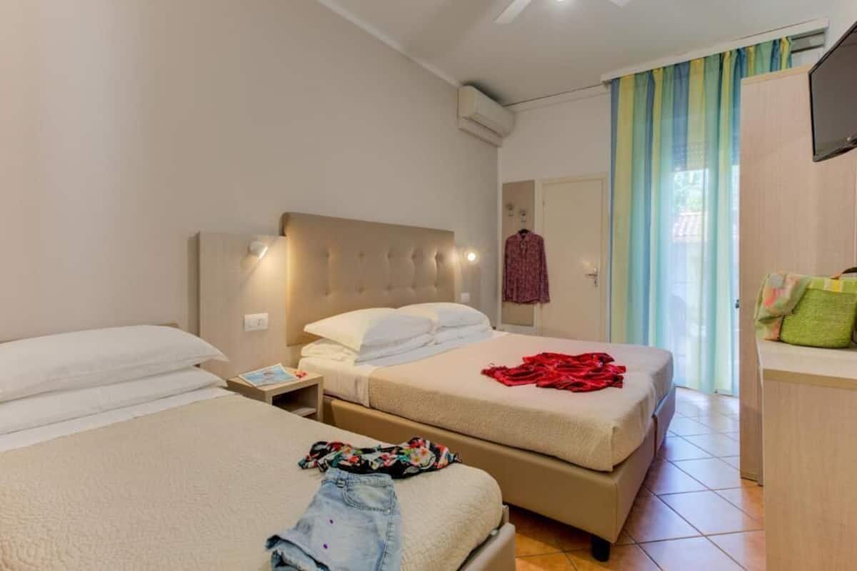 Quarto de hotel com duas camas, um guarda-roupa, uma mesa de cabeceira e uma janela com cortinas.