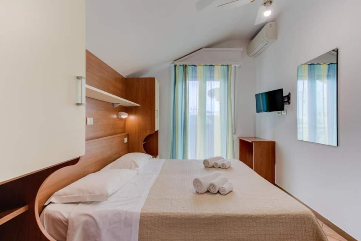 Quarto de hotel com cama, televisão, espelho e janela com cortinas.