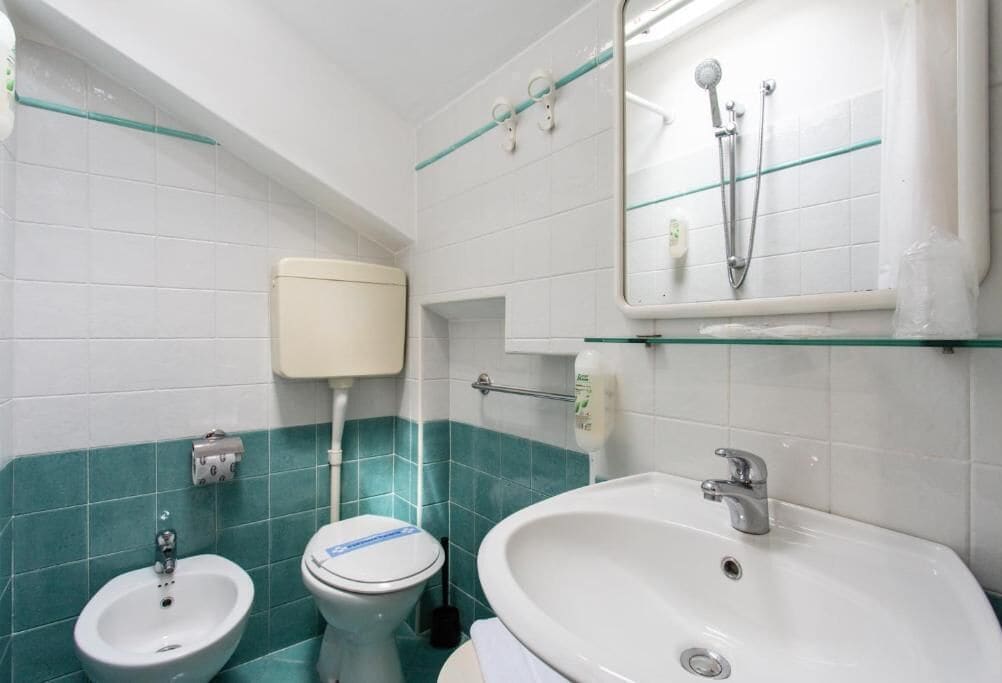 A bathroom with a toilet, bidet, and sink, all with white fixtures and teal tiles.