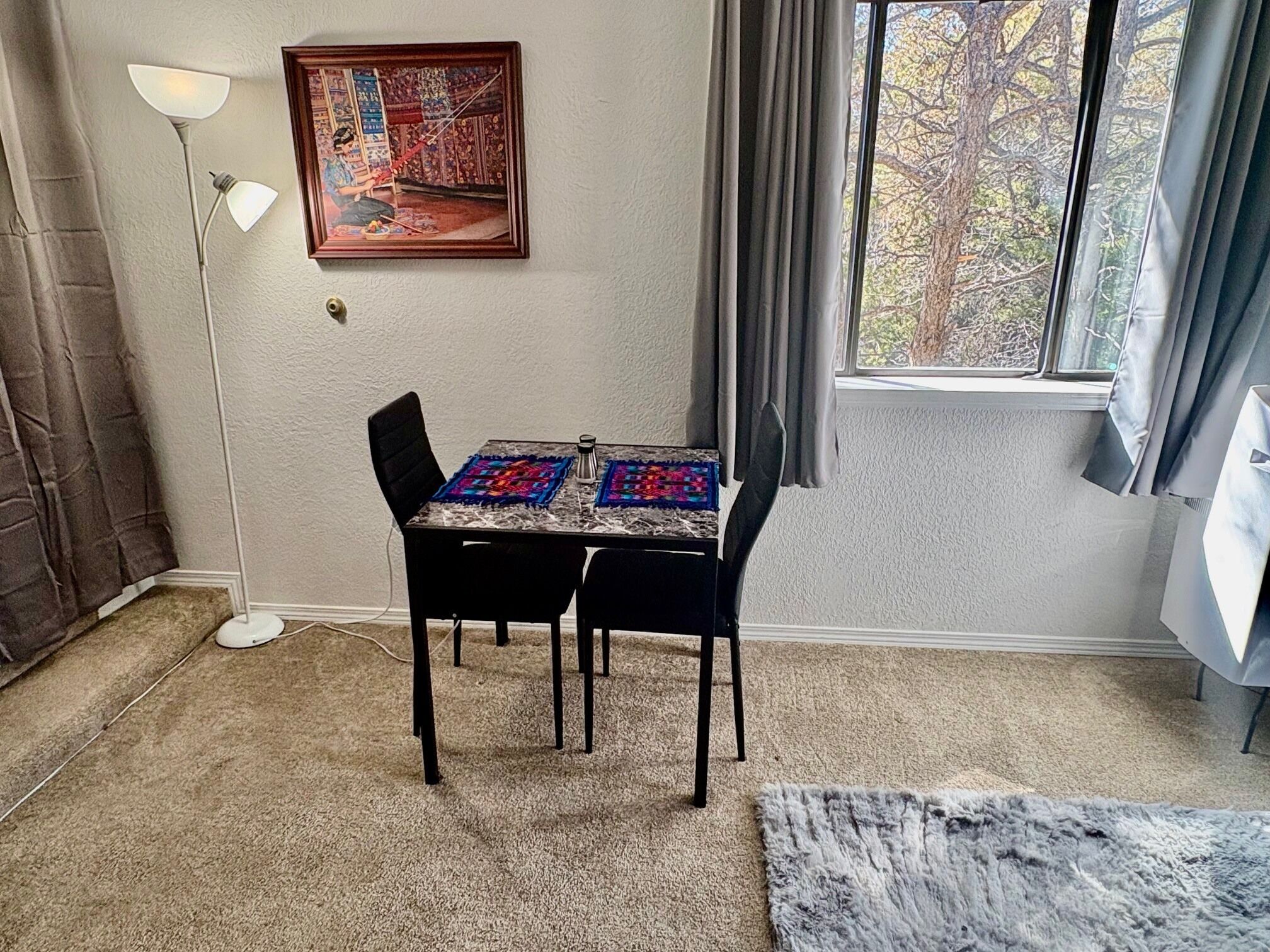 Een kamer met een eettafel, stoelen, een schilderij aan de muur, een raam met gordijnen en een vloerkleed.