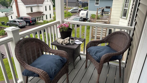 A porch with wicker chairs and a small table, a potted plant, and a view of houses and vehicles in the background. - Newly remodeled cozy 3BR/1BA home on a hill, minutes from town. (Hancock)