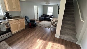A spacious living area with hardwood flooring, a kitchen with a stove and cabinets, a black leather chair, a gray sofa, and a staircase. - Newly remodeled cozy 3BR/1BA home on a hill, minutes from town. (Hancock)