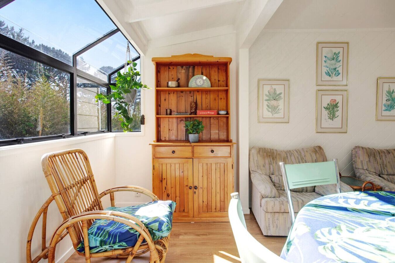 A cozy room with a wooden cabinet, wicker chairs, a sofa, and framed botanical prints on the wall.