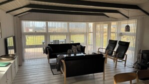 Un salon spacieux avec un canapé noir, deux fauteuils noirs, une table basse en bois et un téléviseur. De grandes fenêtres offrent une vue sur l’extérieur.