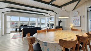 Un vaste espace dédié à la salle à manger, avec une table ronde et des chaises en bois, de grandes fenêtres offrant une vue sur l’extérieur, et un coin salon confortable avec un canapé et des fauteuils.