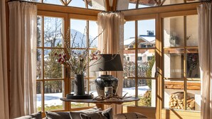 A cozy room with large windows, a wooden table with a vase and branches, and a view of a snowy landscape.