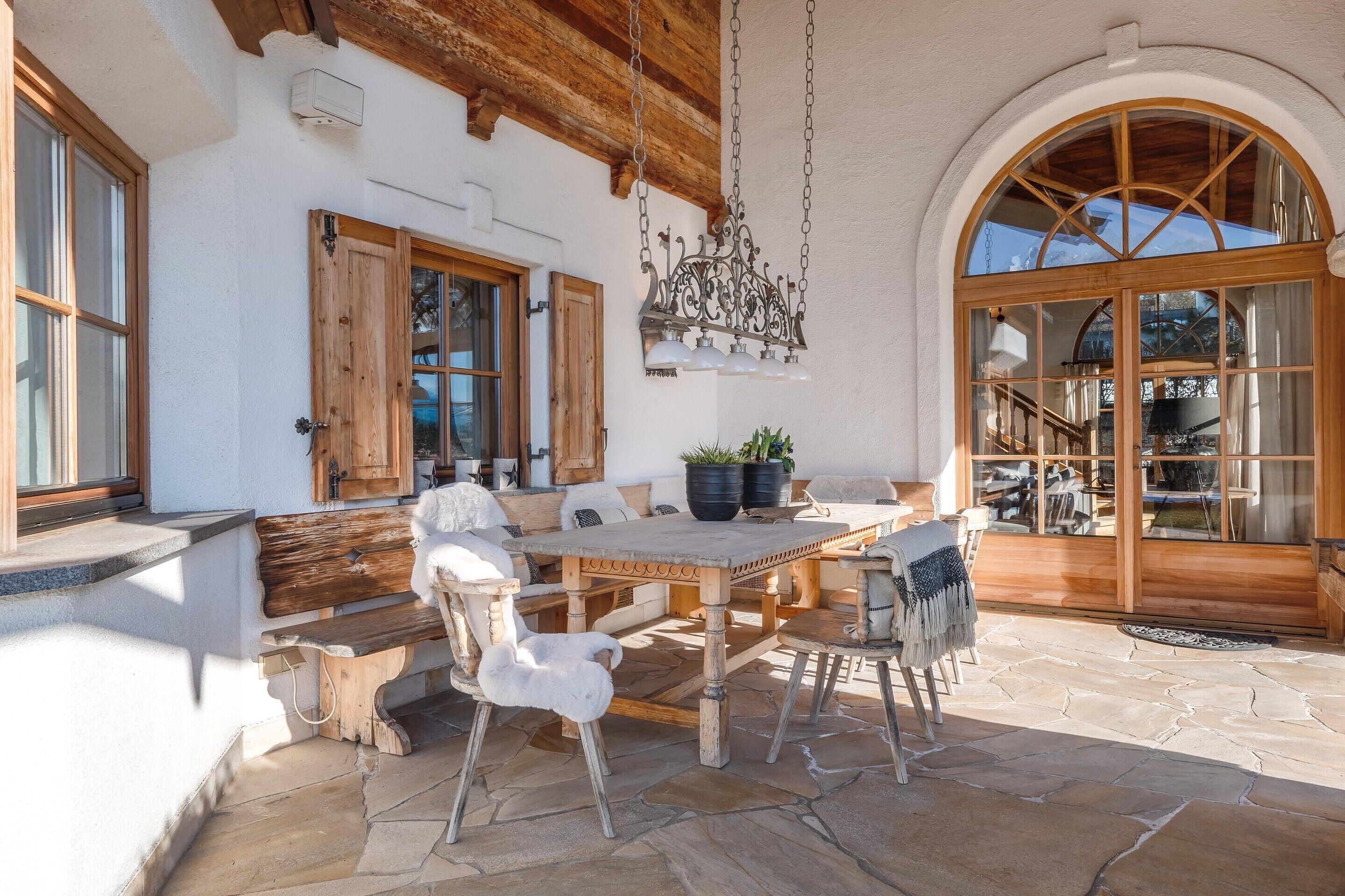 A rustic outdoor dining area with stone flooring, wooden furniture, and a large arched window.