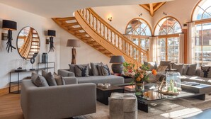 A spacious living room with a wooden staircase, large windows, and modern furniture.