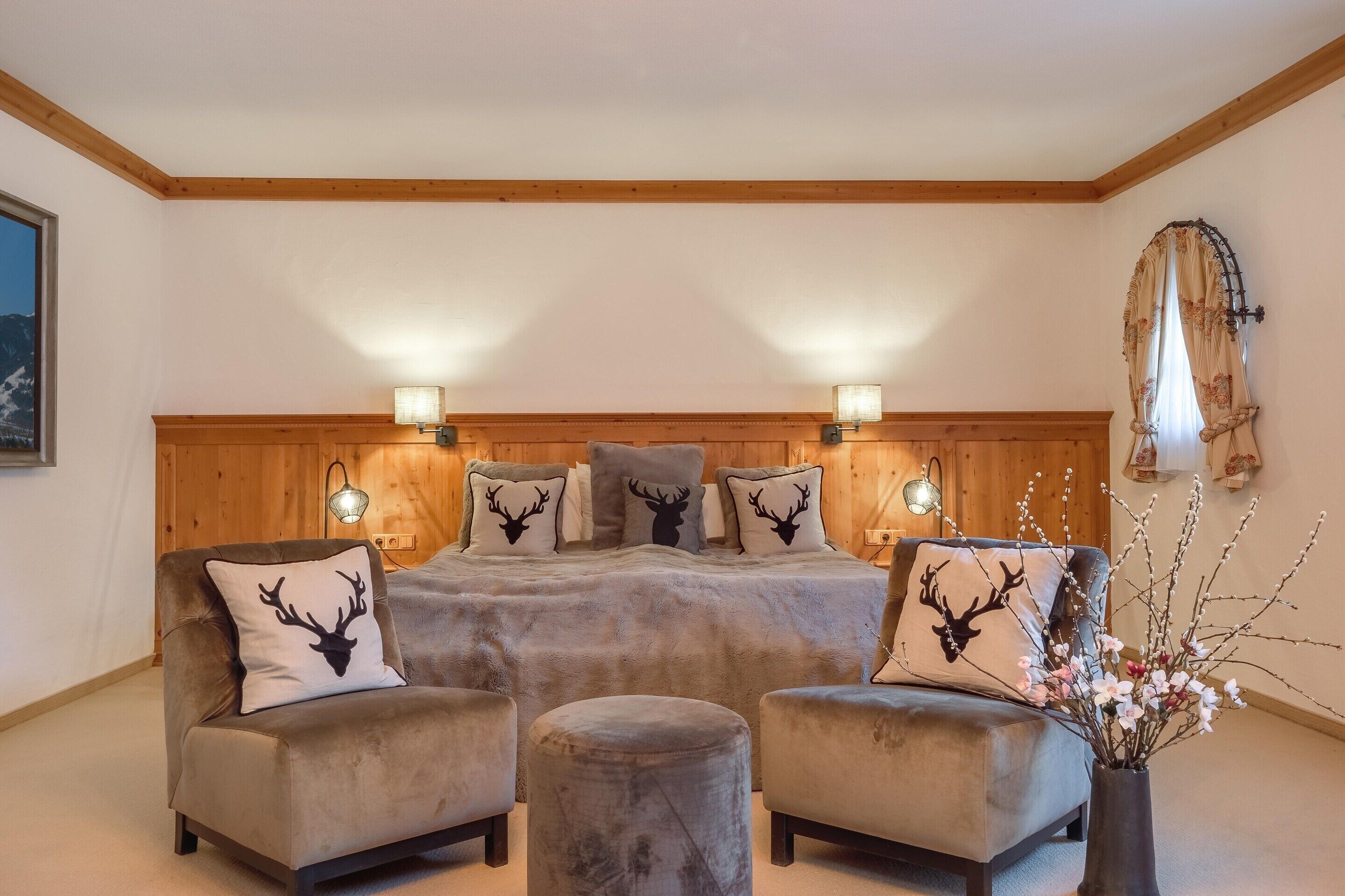 A cozy room with a bed, two armchairs, and a small ottoman. There are wall-mounted lights and a window with a curtain.