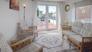 Un salon chaleureux, doté de meubles en osier, d’un tapis à motifs et offrant une vue sur un balcon aménagé avec des sièges d’extérieur.