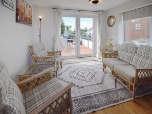 Un salon chaleureux, doté de meubles en osier, d’un tapis à motifs et offrant une vue sur un balcon aménagé avec des sièges d’extérieur.