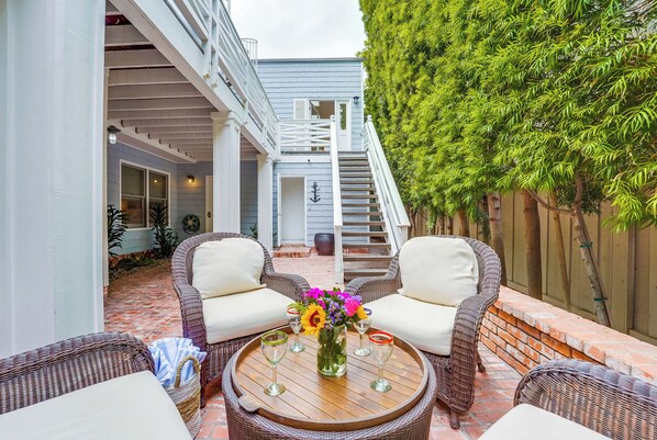 A patio with wicker furniture, a round wooden table, and a staircase leading to an upper level. - The Breakers 1 | 2BR Walk to Windansea Beach (San Diego)