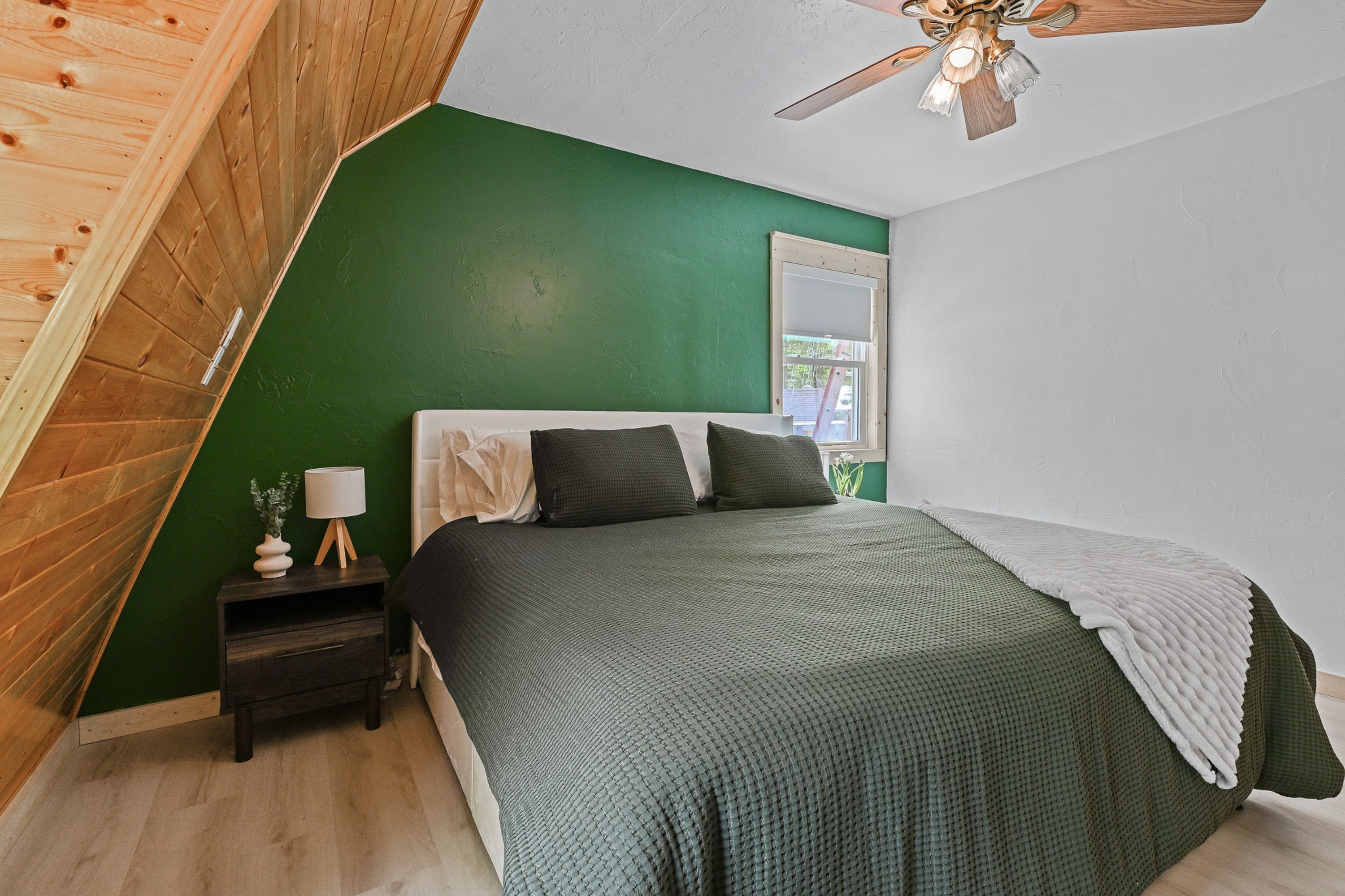 A bedroom with a sloped ceiling, a bed with a green bedspread, a nightstand with a lamp, and a window.