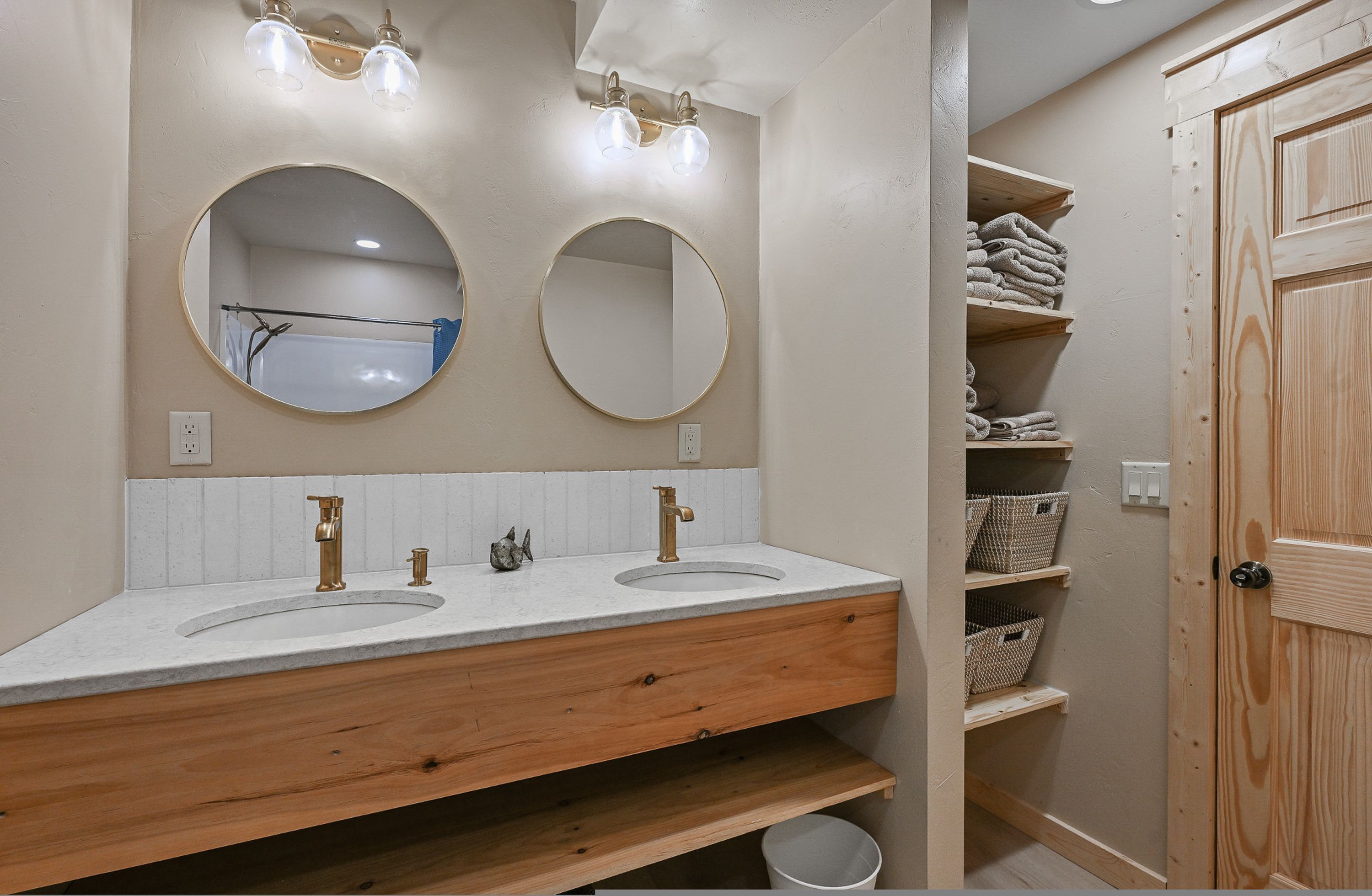 A bathroom with two sinks, wooden vanity, round mirrors, and shelves with towels.