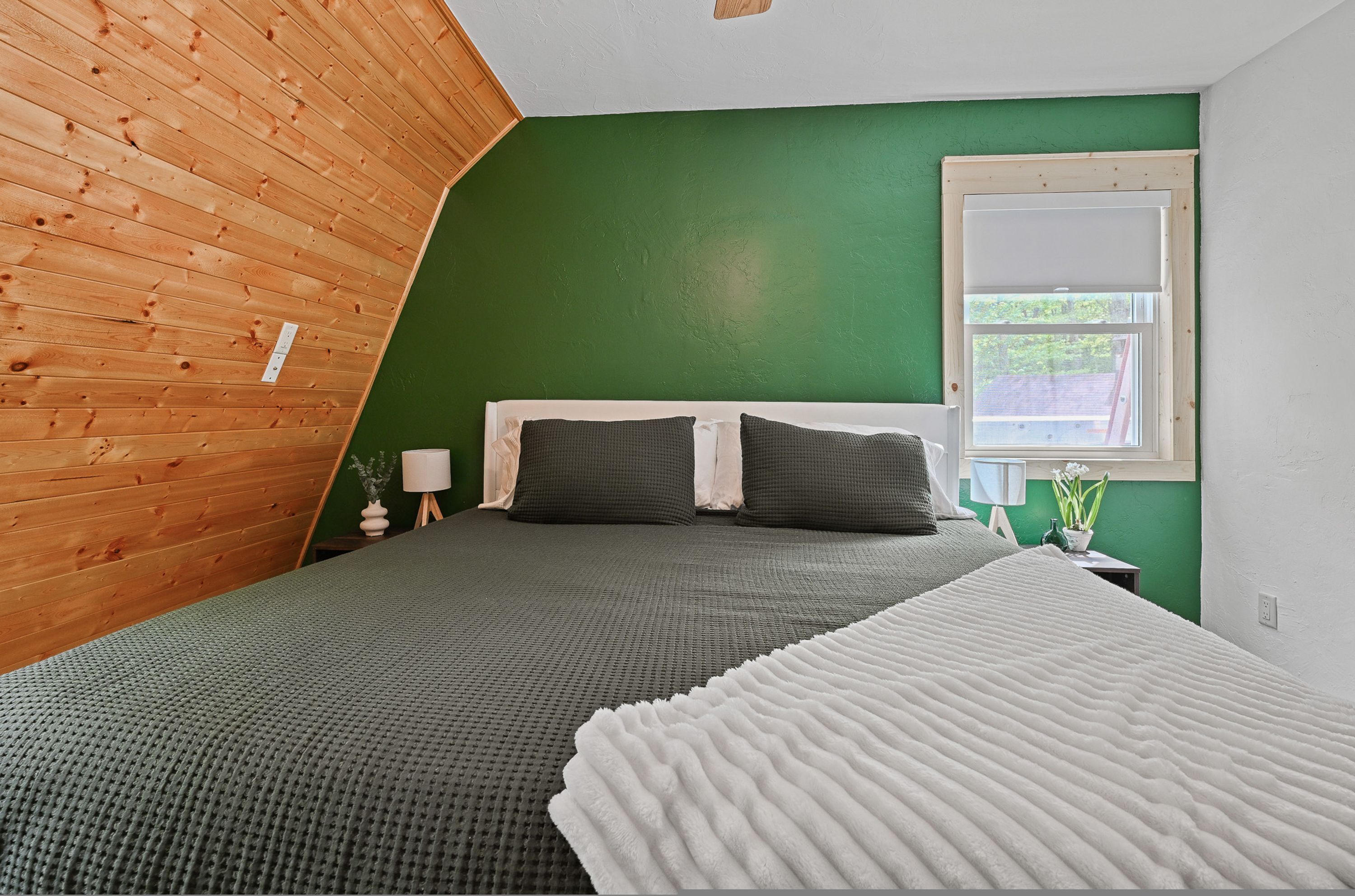 A bedroom with a sloped ceiling, a bed with dark bedding, a white throw blanket, a window with a view of greenery, and a small bedside table with a lamp.