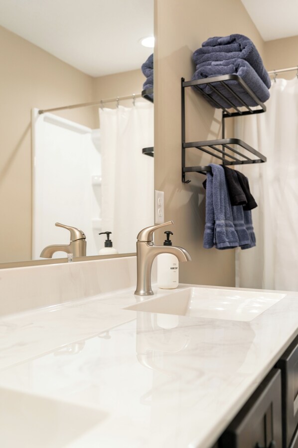 A bathroom with a white sink, two faucets, a towel rack with folded towels, a mirror, and a shower curtain. - Sauna| Garage Golf Games| Close to shopping (Pequot Lakes)