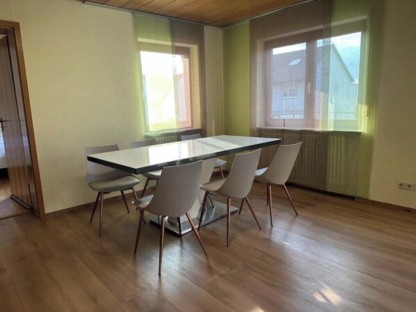 Quarto moderno com mesa de vidro e quatro cadeiras, amplas janelas com cortinas e piso de madeira.