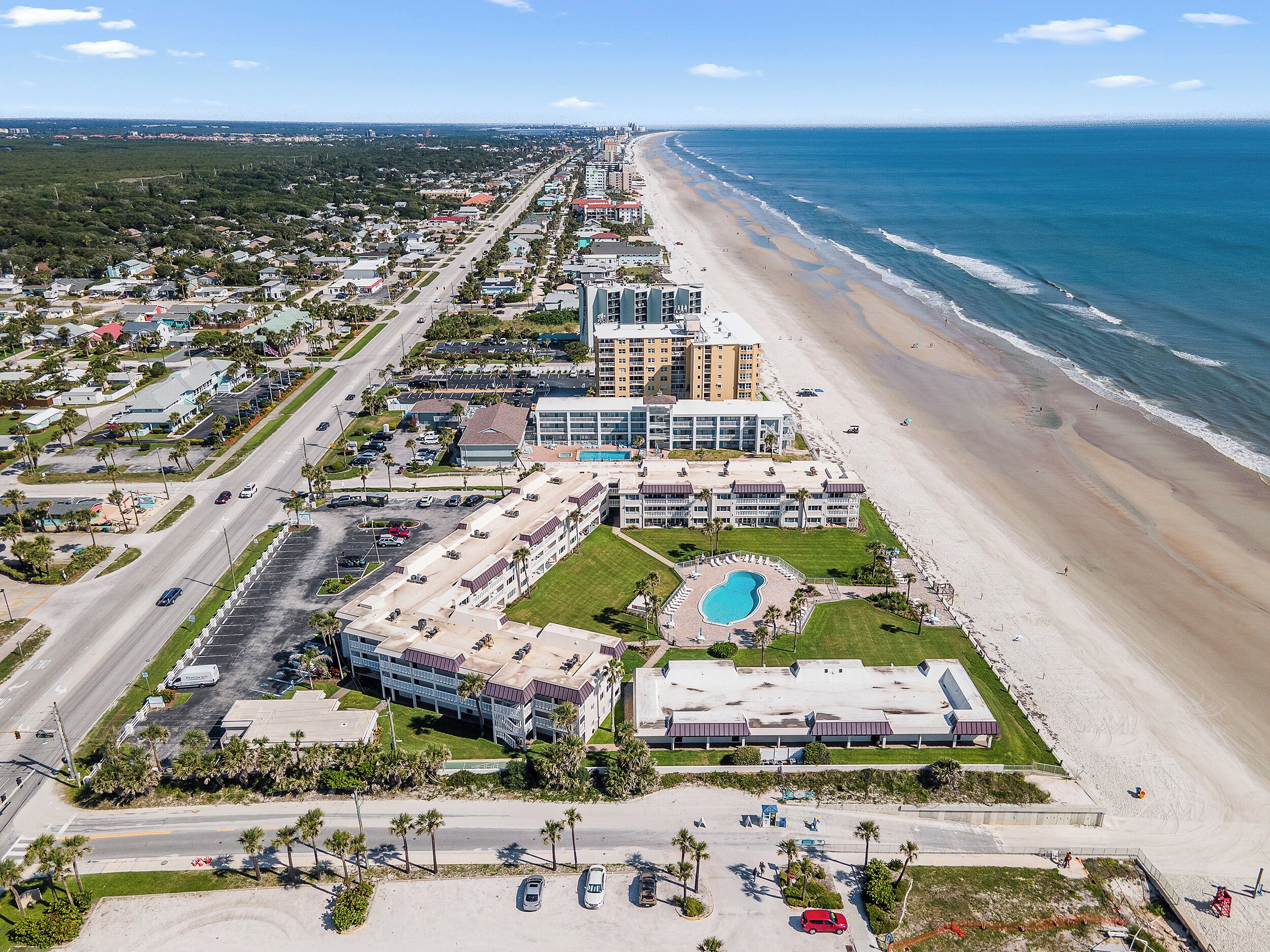 Vue aérienne d’un hôtel situé sur la côte, composé de plusieurs bâtiments, avec une plage et un parking.