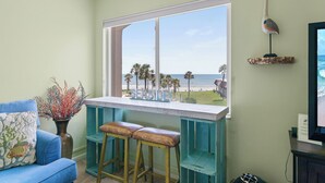 A room with a view of the beach, a blue sofa, a bar area with stools, and a vase with red branches.