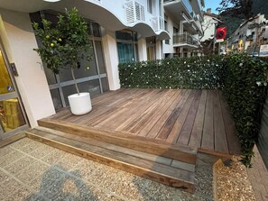 Eine Holterrasse mit Stufen, die zu einem Hauseingang führen, einem Topfbaum und einer Hecke.