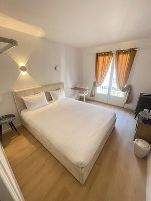 Ein Hotelzimmer mit einem großen Bett, einem kleinen Tisch, einem Stuhl, einem Schreibtisch und einem Fenster mit Vorhängen.