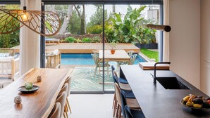 Une cuisine moderne dotée d’une grande fenêtre, un coin repas avec une table et des chaises en bois, et une vue sur une piscine et un jardin.