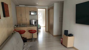 Una sala moderna con un sofá gris, una zona de cocina con armarios de madera, un televisor y un pequeño mueble de madera con un altavoz.
