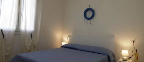 Un dormitorio con una cama, una mesita de noche con lámpara, un aire acondicionado instalado en la pared y una ventana con cortinas.