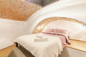 Un dormitori amb un sostre corbat, un llit amb mantell blanc i rosat, i un sòl de fusta.