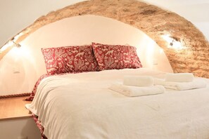 Un llit amb un llençol blanc i dos coixins vermells, una tauleta de nit de fusta i una tovallola blanca.