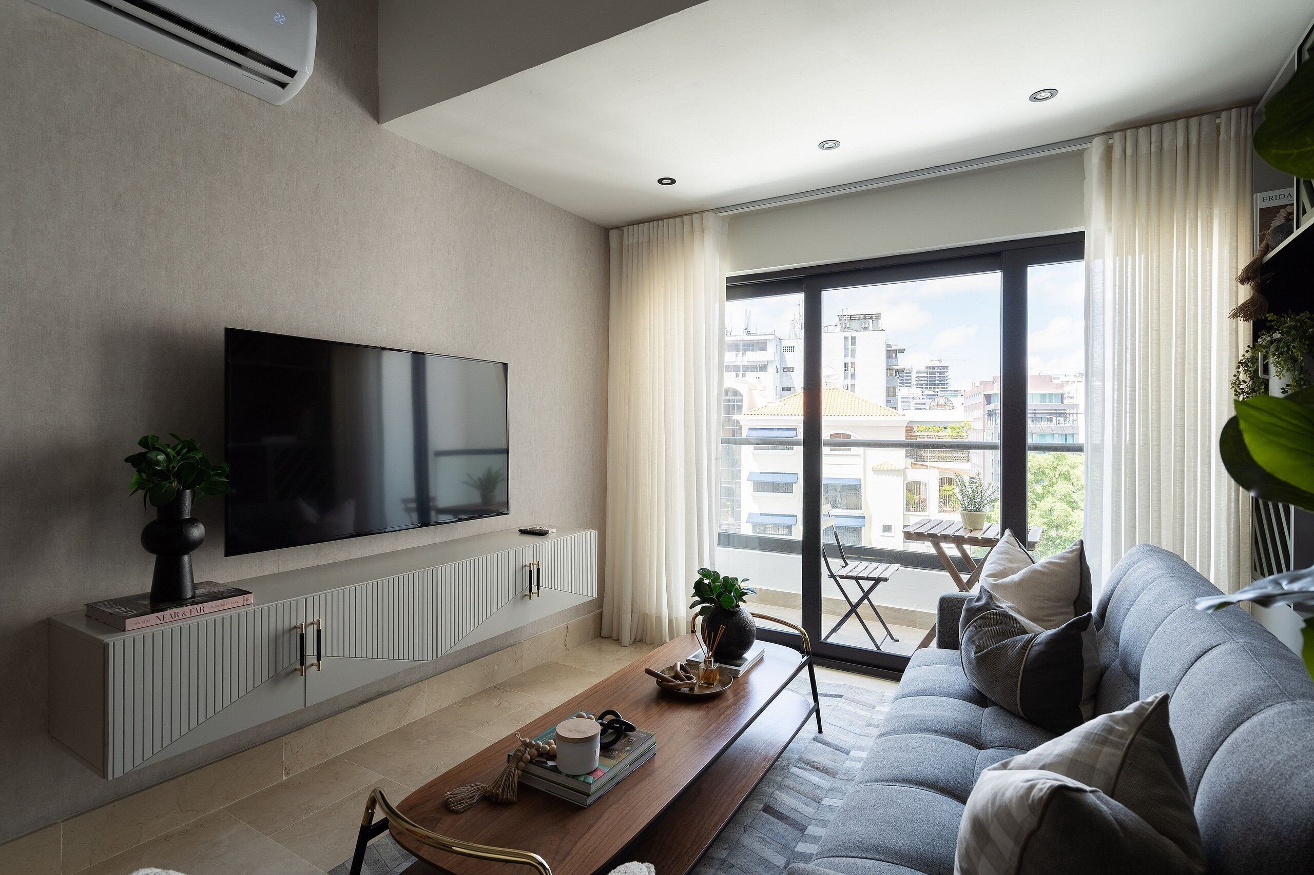 Uma sala de estar moderna com uma TV de tela plana, um sofá, uma mesa de centro e uma janela grande com vista para a cidade.