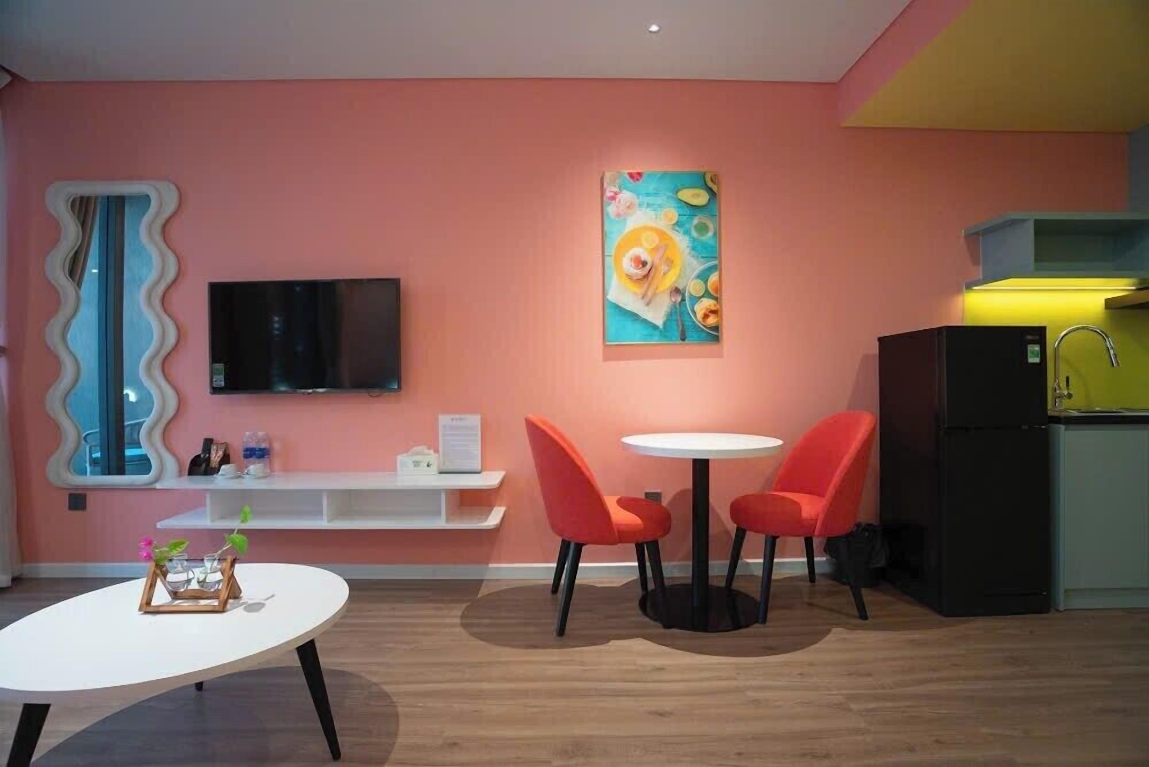 Een moderne woonkamer met een roze muur, een flatscreen televisie, een schilderij, een ronde tafel met twee oranje stoelen en een witte salontafel.
